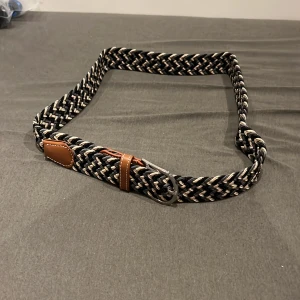 Flätat svart och beige bälte - Snyggt flätat bälte i svart, beige och grått med detaljer i brunt skinn och silverspänne. Bältet har ett coolt mönster och passar perfekt till jeans eller chinos. Flexibel och justerbar längd tack vare det elastiska materialet. Helt ny och oanvänd.
