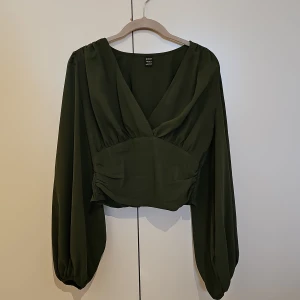 Mörkgrön blus med v-ringning från SHEIN - Snygg mörkgrön blus från SHEIN i storlek S. Blusen har v-ringad omlottfront, långa puffärmar med resår vid ärmslut och smockad midja baktill. Croppad passform som ger en cool siluett. Perfekt att styla med höga jeans eller kjol.