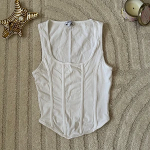 Vit korsett-topp från Divided - Säljer en vit korsett-topp från Divided med markerade sömmar framtill och rundad halsringning. Toppen är ärmlös och har en figurnära passform som ger en snygg siluett. Perfekt att styla med jeans eller kjol för en trendig look.