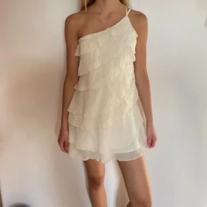 Vit omlottklänning med volanger🤍 - Superfin kort kräm vit klänning med volanger och oneshoulder-design. Klänningen har tunna axelband som går att justera. Bara använd en gång. Perfekt för sommaren! 