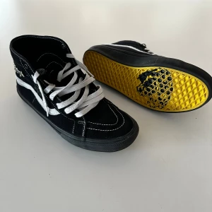Svarta Vans Sk8-Hi med gul sula - Säljer ett par Vans Sk8-Hi sneakers i svart mocka med vita detaljer och snörning. Klassisk vit sidostrip och kontrastsömmar. Yttersulan är gul med svart mönstertryck undertill, vilket ger en unik look. Perfekta för dig som gillar streetstyle och vill sticka ut. 