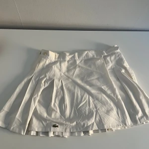 Vit plisserad tenniskjol från Lacoste - Superfin vit plisserad tenniskjol från Lacoste med klassisk krokodillogga framtill. Kjolen är vintage och därav något sliten från användning med diskreta missfärgningar. Strl 42 men mer en S-M, som passar mig med strl 25 i midjan då man kan spänna kjolen med knappar på sidan!
