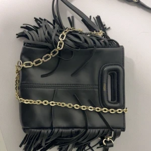 Svart fransad axelväska från Maje - Cool svart axelväska från Maje i skinn med fransdetaljer och guldfärgad kedja. Väskan har ett unikt handtag och trendig design som sticker ut. Perfekt för dig som vill ha en edgy accessoar med lyxig känsla.