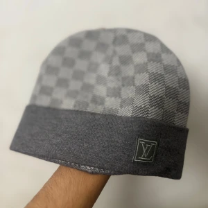 Stilren grå beanie i lyxig design - En snygg och bekväm beanie i grått damier-inspirerat mönster. Perfekt för både vardag och kyligare dagar – kombinerar stil, värme och en exklusiv känsla. 	•	Färg: Grå / Vit 	•	Storlek: One Size 	•	Detalj: Elegant logga framtill 	•	Tidlös, modern design  Passar dig som vill ha en mössa som känns både stilfull och unik. 