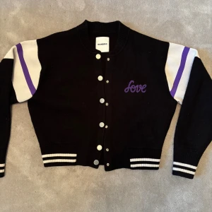 Svart bomberjacka från Sandro med lila detaljer - Svart bomberjacka från Sandro med vita och lila ränder på ärmarna, broderad text 'Love' framtill och stor Sandro-logga på ryggen. Jackan har ribbade muddar, knappar framtill och är tillverkad i en mix av viskos, nylon och polyester. Snygg och sportig collegekänsla.