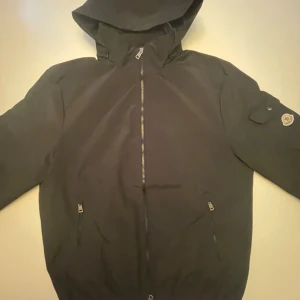 Svart moncler vindjacka  - Snygg och stilren Moncler-jacka i svart. Med fungerande NFC Scan Jackan har en avtagbar huva (fästs med dragkedja) och klassisk Moncler-logga på ärmen. Två praktiska fickor med dragkedja fram och en liten ficka på ärmen. Säljer då den var för stor för mig, men är normal i storleken. Vid frågor samt funderingar så är det bara att fråga!🙂  ✔ Storlek: XL ✔ Färg: Svart ✔ Skick: Som ny – endast provad