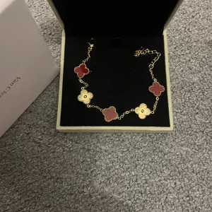 VAN CLEFF armband - Exklusivt Alhambra armband  med ikoniska fyrklöver i guld och röd nyans. Kedjan är tunn och elegant, och armbandet levereras i originalask med äkthetsintyg. Perfekt statement piece för dig som gillar lyxiga detaljer.