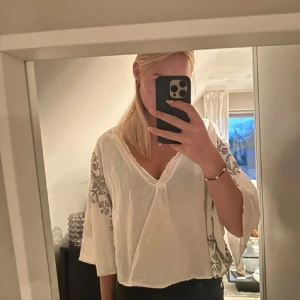 Vit blus med broderi från Zara - Luftig vit blus från Zara med v-ringning och spetskant. Vida trekvartsärmar med snyggt svart broderat mönster längs ärmarna. Perfekt till jeans eller kjol för en chill och bohemisk vibe.