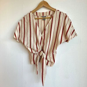 Randig omlottopp med knyt - Kortärmad omlottopp från LINDEX i beige och röd med vertikala ränder. Toppen har v-ringning och knyt framtill, vilket ger en avslappnad och trendig look. Tillverkad i ett lätt, strukturerat tyg som känns luftigt och passar perfekt till varmare dagar.