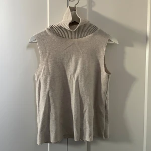 Beige ärmlös polotopp - Säljer en beige ärmlös polotopp med hög, ribbad krage. Toppen är stickad och har en mjuk, avslappnad passform! 