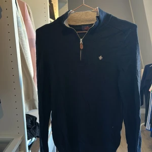 Mörkblå merinoullströja Morris M - Snygg mörkblå stickad tröja från Morris i 100% merinoull. Tröjan har half zip med läderdetalj och en liten broderad logga på bröstet. Perfekt för dig som gillar klassisk och stilren look med lång ärm och hög krage.