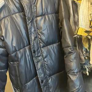 Svart pufferjacka med huva - Säljer en svart pufferjacka med huva och dragkedja framtill. Jackan har stora quiltade partier, sidofickor med dragkedja och tryckknappar längs öppningen. Perfekt för kalla dagar och riktigt skön att bära. Den har använt några gånger men är dock på en ganska bra skick