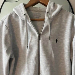 Ralph Lauren zip - Säljer en stilren ljusgrå hoodie från Ralph Lauren med dragkedja och huva. Tröjan har snörning i huvan och den klassiska svarta Polo-loggan broderad på bröstet. Perfekt för en avslappnad look. Jätte fint skick och inga slitningar förutom att de är lite nopprigt vid brösted men syns knappt, skriv privat om du har några funderingar! Mvh