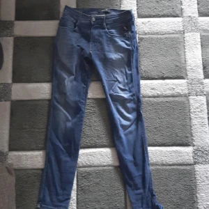 Replay Anbass blå jeans herr - Snygga blå Replay Anbass jeans med klassisk femficksdesign och diskreta slitningar. Jeansen har normal passform och raka ben, med Replay-logga på fickan och metallknapp framtill. Perfekta för dig som gillar stilrena och bekväma jeans.