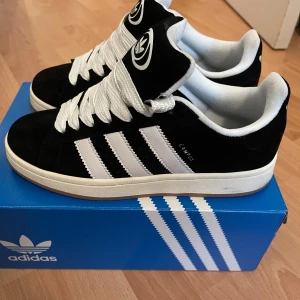 Adidas Campus 00s svart/vit sneakers - Adidas Campus 00s sneakers i svart mocka med vita detaljer och klassiska tre ränder på sidorna. Tjocka vita skosnören, vit sula och Adidas logga på plös och häl. Stilren och retro vibe, perfekt för dig som gillar streetwear.