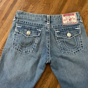 True Religion jeans - Ett par blå jeans från True Religion med låg midja och bootcut-stil. Jeansen har dekorativa sömmar och knappar på bakfickorna (AI). Säljer då de tyvärr är för stora för mig. skriv vid frågor!