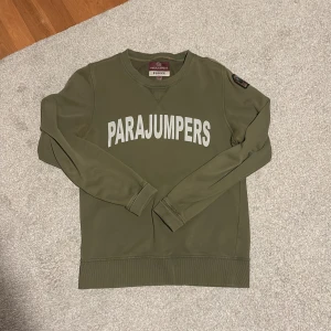 Olivgrön fleece tröja från Parajumpers - En bekväm olivgrön fleece tröja från Parajumpers med texten 'PARAJUMPERS' tryckt i vitt på framsidan. Tröjan har långa ärmar och ribbstickade muddar vid ärmslut och nederkant. Det finns en broderad patch på vänster ärm. Tröjan är i bra skick och har bevarats väl. Nytt pris: 2300kr! Pris kan självklart diskuteras !