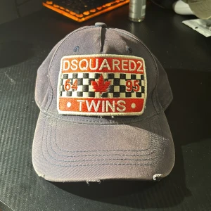 Mörkblå keps från Dsquared2 - En mörkblå keps från Dsquared2 med ett broderat märke på framsidan som visar texten 'DSQUARED2 TWINS' och en röd lönnlöv. Kepsen har en justerbar rem på baksidan och texten 'DEAN & DAN CATEN' broderad i rött. Riv märkerna på framsidan av kepsen är en del av designen. Pris kan diskuteras! 