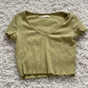 Grön ribbad topp från Pull&Bear - En grön ribbad topp från Pull&Bear med korta ärmar och v-ringning. Toppen har en croppad design med vågiga kanter vid ärmslut och nederkant.