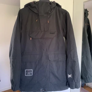 L1 Anorak Skidjacka  - Svart oversized anorakjacka med huva och en ficka på bröstet. Det finns även fler fickor för förvaring. Jackan är använd som skidjacka, men har inga tydliga tecken på slitage. 