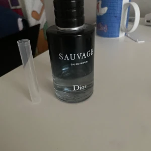 Sauvage Eau de Parfum från Dior - En flaska Sauvage Eau de Parfum från Dior. Flaskan är cylindrisk med en svart och genomskinlig design, där den övre delen är svart och den nedre delen är genomskinlig. Parfymen har en elegant och modern look.