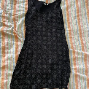 Svart klänning från Sinecura - En svart ärmlös klänning från Sinecura med ett cirkulärt mönster i vitt. Klänningen har en rund halsringning och är i storlek S.