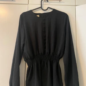 Svart blus - En svart slät blus med en dragkedja bakom ryggen och små detaljer från bröstet till midjan. Den är mellan lång blus som sitter vid midjan och som är löst nedåt