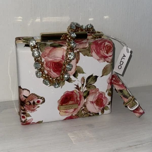 Blommig Aldo väska  - Blommig Aldo väska. Helt ny, köpt från USA för 45$ som motsvarar ca 480kr, jag säljer den nu för 440kr. 🌸🩷 ca 12cm höjd och 14 cm längd. Man kan ha den kort men också med det lång bandet. Satin tyg inuti. Helt felfri! 🩷🌸