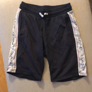 Svarta shorts från Gant - Svarta shorts från Gant med vita sidopaneler där märkesnamnet 'GANT' är tryckt flera gånger. Shortsen har en elastisk midja med dragsko för justering och två sidofickor.