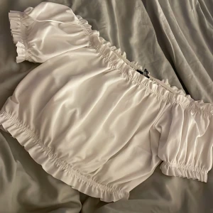 Vit off-shoulder topp - En vit off-shoulder topp med korta ärmar och elastiska band vid axlar, ärmslut och nederkant. Toppen har en lös passform och är tillverkad i ett lätt och bekvämt material.