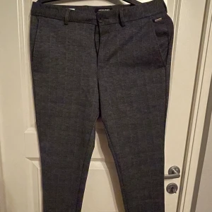 Kostymbyxor  - Rutiga kostymbyxor i mörkgrått. Två fickor framtill samt två fickor baktill, försluts med dragkedja och knapp.