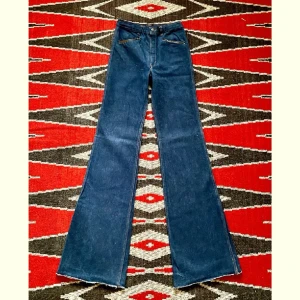 70-tal, Las Vegas jeans - Asballa jeans med läderdetaljer av Las Vegas på 70-talet. Bra skick, några stygn saknas på höger bakficka och färgen har blött in på mocka-lappen i bak.  Midja 34cm Höfter 43cm Gren 28cm Längd 120cm Innersöm 92cm Ofållade byxben  100% bomull
