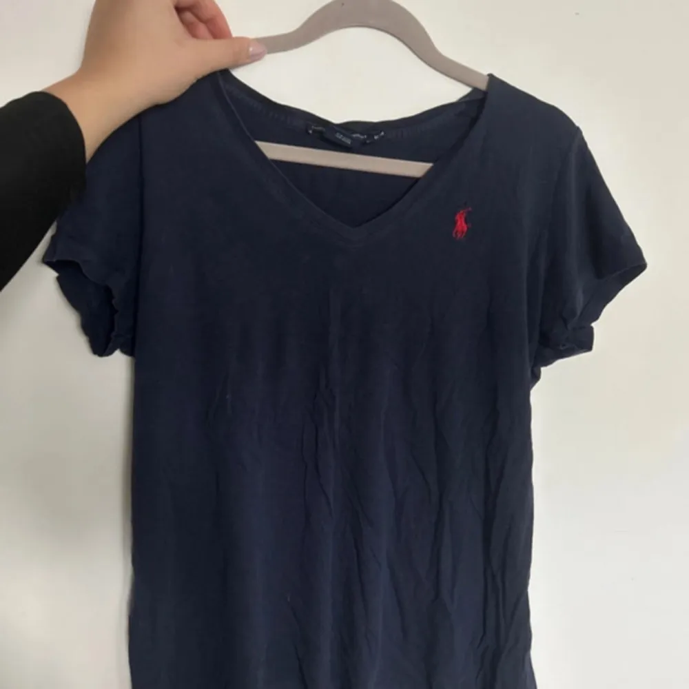 En mörkblå t-shirt från Ralph Lauren Sport i storlek M. T-shirten är tillverkad av 100% bomull och har en röd broderad logotyp på bröstet. Den har en v-ringad hals och korta ärmar. Har tre lagade hål . T-shirts.