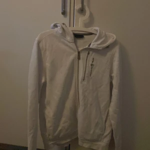 Vit hoodie från Sail Racing - En vit hoodie från Sail Racing med dragkedja framtill. Tröjan har en huva med ett litet tryck på baksidan och en dragkedja på bröstet. Den har någon fläck på framsidan men ska försöka tvätta bort det!