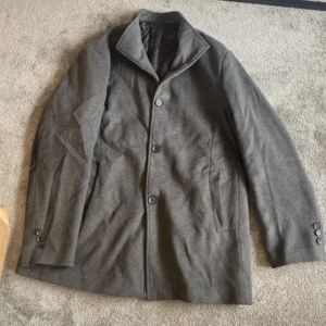 Grå rock - En stilren grå rock med knappar framtill och på ärmsluten. Rocken har en klassisk design med en krage och är tillverkad i ett mjukt material. En knapp (se bild) har gått sönder. Annars är kappan endast använd 1 gång 
