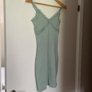 Grön klänning från H&M Divided - En grön klänning från H&M Divided i storlek XS. Klänningen har spetsdetaljer och en liten rosett framtill. Den är ärmlös och har en tight passform.