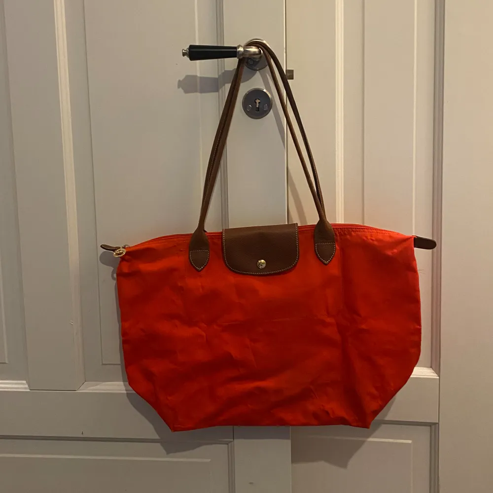 En snygg Loncchamp väska i knall orange färg. Modell Lapliage Väskan har tecken på användning där av priset. Läng:48 cm Bredd:32cm. Den har ett litet hål (se bild 5) och lite missfärgad. Hör av er vid frågor eller funderingar! 🧡🟠. Väskor.