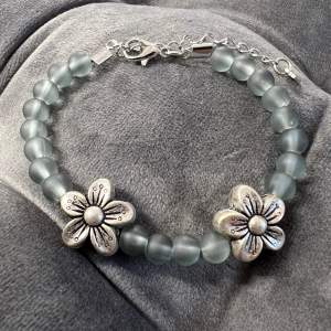 Handgjort pärlarmband i gråa pärlor med två fina silverfärgade blommor, även spännet är silverfärgat. Justerbar passform mellan 17-22 cm.