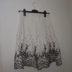 Blommig a-linjekjol i svart och vitt - En söt midrise a-linjekjol med spetsliknande blommönster längst sömmen i svart och gräddvitt. Kjolen har öven en underkjol.