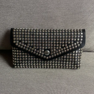 Mobilväska  - Mobilväska från Don Donna. Finns hållare på baksidan för att kunna sätta den på ett skärp. I svart syntet med platta nitar. Aldrig använd.  Mått: bredd 18cm, höjd 10cm, djup 3cm. 