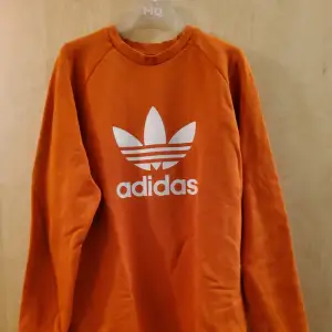 Härligt orange, med vitt tryck, collegetröja från Adidas. Oversize modell.  Material: bomull och elastan