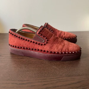 Louboutin Slip-ons - Skitcoola vinröda Louboutin slip-ons med alligator mönstrad mocka. Ibga taggar har lossnat och skorna är i fint skick! Storlek 42 passar även 41! Inget pg ingår.