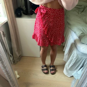 Kjol med små blommor - Fin röd kjol med små vita blommor på med en Dido volang och rosett i sidan. Dragkedja bak. Storlek S