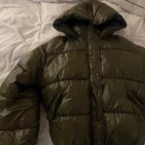 Grön glansig pufferjacka - Storlek xs/S men sjukt stor. Passar snyggt oversized på en M! Cool detalj med dragkedja på luva. Sjukt fluffig och skön