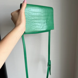 Liten handväska  - Liten handväska från Carin Wester i en härlig grön färg. Använd men i bra skick. Smalt och brett band ingår (det breda är helt oanvänt) Obs! Trasig vid öppningsfacket, se sista bilden, men ingenting som syns såvida väskan inte är upp och ned