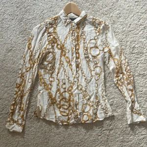 Vit skjorta med kedjemönster från Zara Woman - En elegant vit skjorta/blus i 100% siden från Zara Woman med ett unikt kedjemönster i guld. Skjortan har långa ärmar och knappar framtill. Perfekt för både vardag och festliga tillfällen. knappar i äkta pärlemor. nedersta knappen saknas men ersätts lätt av standardknapp