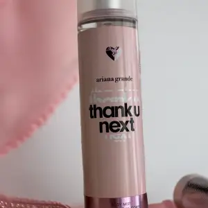 En rosa body mist från Ariana Grande med namnet 'Thank U Next'. Flaskan har en hjärtformad logotyp och en genomskinlig topp. Produkten innehåller alkohol och parfym.