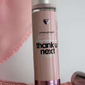 Thank U Next Body Mist från Ariana Grande - En rosa body mist från Ariana Grande med namnet 'Thank U Next'. Flaskan har en hjärtformad logotyp och en genomskinlig topp. Produkten innehåller alkohol och parfym.