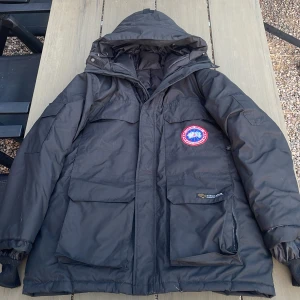 Svart dunjacka från Canada Goose - En svart dunjacka från Canada Goose med huva och flera fickor. Jackan har en broderad logotyp på bröstet och en etikett på ryggen. Den är tillverkad i Kanada och är avsedd för kalla klimat.
