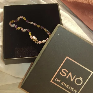 Snö of Sweden armband  - Ett elegant armband från SNÖ of Sweden med färgglada stenar i lila, gult, rosa och vitt. Armbandet har en guldfärgad bas och ett säkert lås. användt cirka två gånger 
